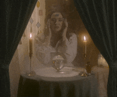 Video gif. Woman dressed as a fortune teller sits at a circular table with candles and tarot cards. She rubs her forehead in concentration over crystal ball and text appears, "I see it so clearly." She slowly opens her eyes making a wild expression at us. 