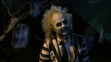 Movie gif. Michael Keaton as Beetlejuice sits in a graveyard while the lightning flashes. He holds his arms out and smiles as he says, “It’s showtime.” 