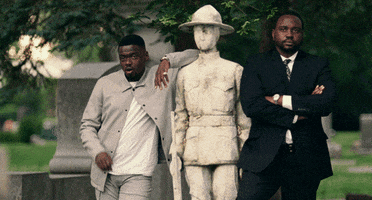 Movie gif. Daniel Kaluuya as Jatemme and Brian Tyree Henry as Jamal Manning stand next to a statue of a soldier. Jatemme leans on the statue's shoulder and gives a small wave as he chews on gum. Jamal has his arms crossed and has a serious look on his face.
