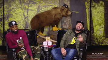 Video gif. Desus and Mero sit next to each other, behind them a taxidermy bear wearing a hat and yellow boots. Mero leans back with a cheeky smile on his face and moves his arms around to demonstrate something. Desus leans forward with a slight smile on his face, listening.