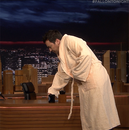 Late Night gif. Jimmy Fallon wears a fancy robe and picks up his mug from his desk. He walks around, sipping from his mug, squinting one of his eyes.