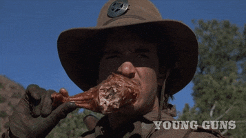 Movie gif. Dermot Mulroney as Dirty Steve Stevens in Young Guns. He's squinting at something far in the distance and as he gnaws on a big turkey leg.