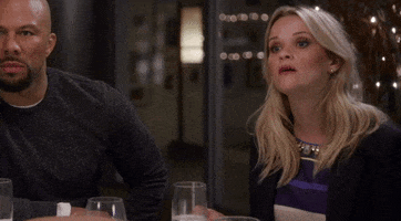 Reality TV gif. Reese Witherspoon on the Chelsea Handler show sits at a dining room table next to Common. Reese’s eyes get big and she cringes in shock. Common nods and then pauses in surprise at the same time Reese cringes. 