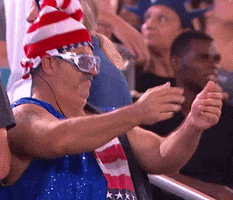 Video gif. An older man dressed in an American flag tank top, sparkly blue vest, shutter sunglasses, and an American flag hat that’s shaped like a hand doing the peace sign. He claps while doing a little dance, and then looks over at us and points. 