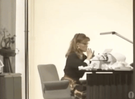 Video gif. Vintage video of a woman sitting at a typewriter, holding her chin in her hand. The camera zooms in and she tosses her hands in the air before dropping her head into her hands in frustration.