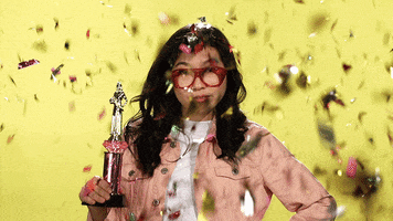 Celebrity gif. Awkwafina wears red glasses on her face while she stands with her hand on her hip. In her other hand is a golden trophy. Golden confetti falls down around her and catches in her hair. She stares at us with a smug look. 