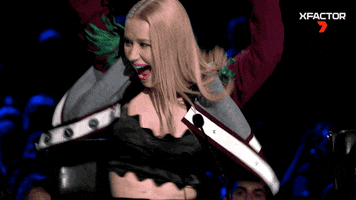 Reality TV gif. Iggy Azalea on X Factor shakes wildly as she sits at the judges desk. She raises her arms up in the air and shakes her head, smiling.