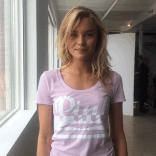 Video gif. Woman smiles at us and waves cutely as she wears a Victoria’s Secret Pink t-shirt.