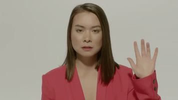 Music video gif. Closeup of Mitski with a dazed expression as she waves at us in her video for Your Best American Girl.