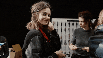 Video gif. Woman at New York Fashion Week looks at us and waves, smiling, her phone in her hand.
