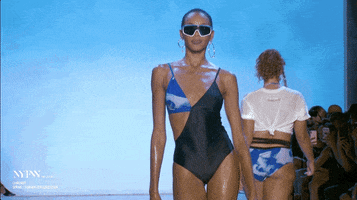 Video gif. Model struts down the runway in a swimming suit and dark sunglasses. She then waves her hand to fan herself like she’s too hot.
