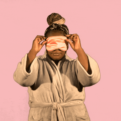 Video gif. Against a soft pink background, a woman in a beige bathrobe with dreadlocks in a bun lifts a shiny pink sleeping mask from her eyes. She reacts with excitement as she notices us, and greets us with a big smile. Yellow text grows from bottom of frame, "Good morning."
