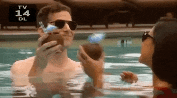 TV gif. Andy Samberg as Jake Peralta and Melissa Fumero as Amy Santiago in Brooklyn 99 toast with two tropical coconut drinks while floating in a pool. 
