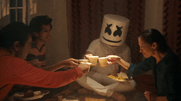 Music video gif. Marshmello sits on the floor with a family and they all cheers together, drinking tea and eating dinner.