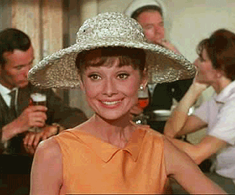Celebrity gif. Audrey Hepburn sitting at a cafe and sees someone she recognizes. She gives a nervous grin and waves hi in a circle motion.