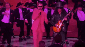 Oscars 2024 GIF. Ryan Gosling, dressed in a Barbie pink tuxedo and sunglasses, clutches the air with drama, flanked by Slash and a chorus line of dancing Kens.