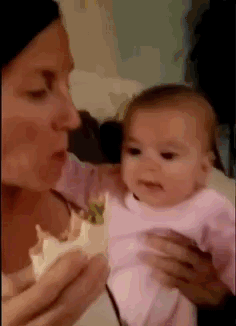 Video gif. A baby sits in the lap of a woman holding a burrito. The baby opens its mouth wide and leans in for a big bite as the woman holds the burrito out of reach.