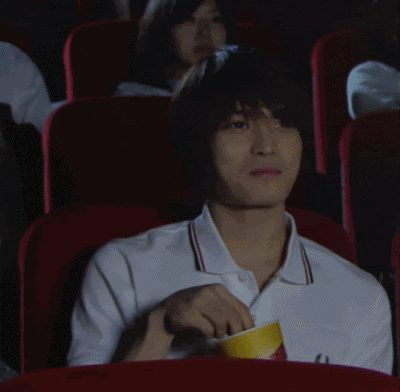 Celebrity gif. Jaejoong smiles while eating popcorn, sitting in a red movie theater seat.