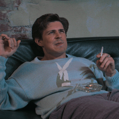 TV gif. Chris Lowell as Bash Howard in GLOW relaxing on a couch and smoking. He's smiling, but his face contorts into a look of concern as if coming to a sudden realization.
