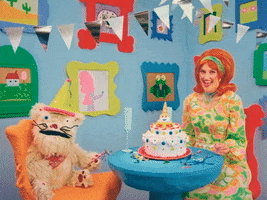Video gif. Woman wearing a 50s-style orange wig smiles sweetly at a furry puppet across the table then points at us. The room they are sitting in is decked out with a cake on the table and bright blue walls filled with cute, colorful framed illustrations of animals, food, and scenes. Text below says, "Happy birthday, you old fart!" Then the puppet says, "Who's she calling old fart?'