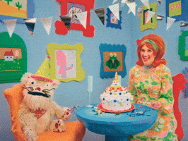 Video gif. Woman wearing a 50s-style orange wig smiles sweetly at a furry puppet across the table then points at us. The room they are sitting in is decked out with a cake on the table and bright blue walls filled with cute, colorful framed illustrations of animals, food, and scenes. Text below says, "Happy birthday, you old fart!" Then the puppet says, "Who's she calling old fart?'