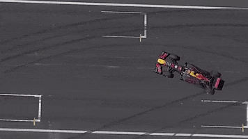 Video gif. Drone footage looks down on a formula one car making donuts on a blacktop. Smoke billows out from the back tires.