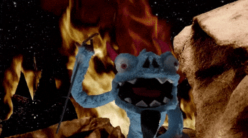 Video gif. Blue devil puppet screams at us and waves around a red pitchfork as he stand in front of rocks and raging fire.
