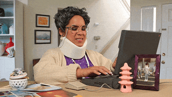 Video gif. Elderly woman has a neck brace on and she slowly types away at an antiquated laptop. She moves slowly as she types and squints her eyes at the screen.