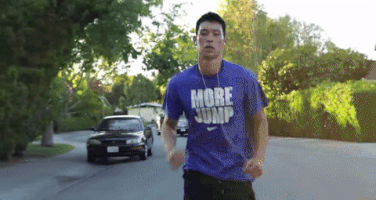 Sports gif. Jeremy Lin sweats as he runs down the middle of a residential street lined with green shrubs and leafy trees.