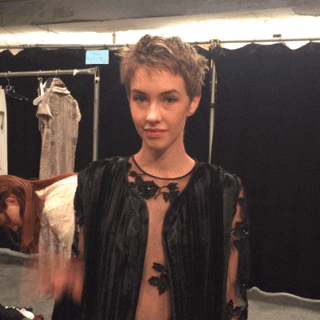 Video gif. Short-haired model in a fancy black and sheer shirt waves at us with a smile and mouths "hi".