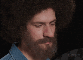 Video gif. Man dressed like Bob Ross has a sad look on his face as he looks down. He then slowly raises his head up and as he does, a smile spreads across his face and he looks around like he realizes everything is beautiful.