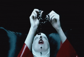 Music video gif. From the video for Lady Gaga's Alejandro, a nun leans against a black statin pillow and carefully feeds a rosary down her throat.