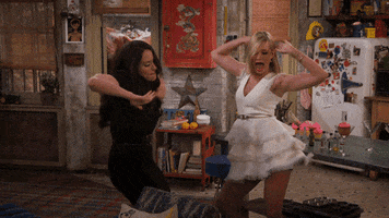 TV gif. Kat Dennings as Max and Beth Behrs as Caroline in Two Broke Girls dance with abandon in a cluttered apartment.