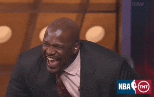 Sports gif. Wearing a dark gray suit jacket, Shaquille O'Neal leans forward in his seat for a prolonged, emphatic laugh.