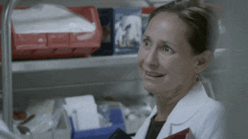 TV gif. Laurie Metcalf as Jenna on Getting On, standing near a shelf holding medical supplies, with a straight face that morphs into a strained smile as she says, "great!" which appears as text.
