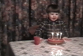 Video gif. Old home video of a child headbanging while blowing out three birthday candles on a cake.