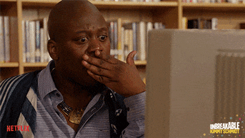 TV gif. Tituss Burgess as Titus Andromedon from Unbreakable Kimmy Schmidt covers his mouth in a shocked reaction to something on the screen of a library computer.