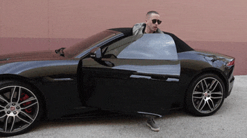 Celebrity gif. Wearing sunglasses and glancing around, Yandel slowly emerges from a black Jaguar convertible.