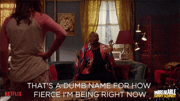 TV gif. Tituss Burgess as Titus in Kimmy Schmidt rises from a sofa in a colorful robe and puts his hand on his hip, serving looks, while saying, "That's a dumb name for how fierce I'm being right now," which appears as text.