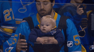 Sports gif. St. Louis Blues hockey fan wearing a jersey sits in the crowd as his baby, strapped to his chest in a baby carrier, stares blankly.