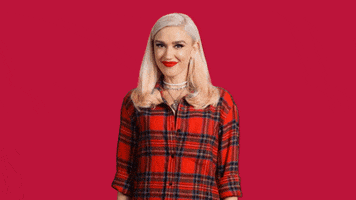 Celebrity gif. Gwen Stefani, wearing a red plaid shirt and standing in front of a red background, smiles at us and waves her hand.