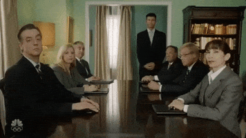 SNL gif. The cast of SNL are all at a large table and sit in suits with their hands folded. They stare at us intimidatingly as they lean in to stare at us harder.