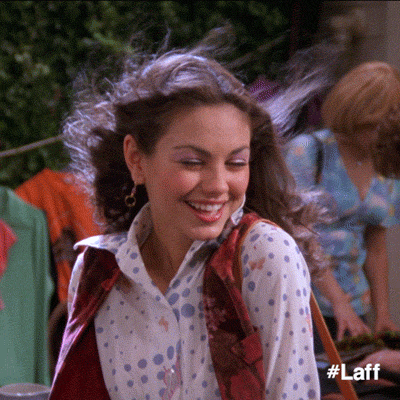 TV show gif. Mila Kunis as Jackie in That 70's Show laughs as the wind blows her hair. She places her palm on her chest, then composes herself with a flirty glance.