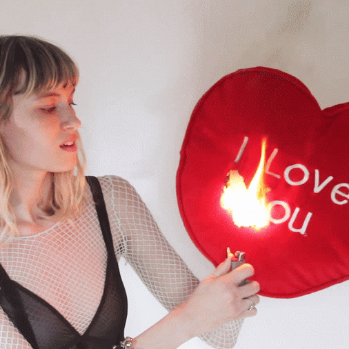 Video gif. Woman is holding a red heart pillow that says, "I love you," and she's lighting it on fire.