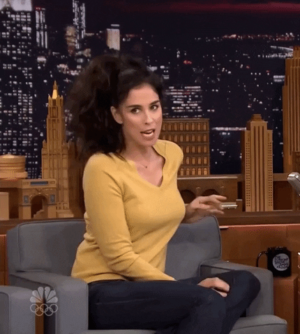 Tonight Show gif. Sarah Silverman sits on the gray sofa wearing a yellow V-neck sweater and her hair tied into a casual ponytail. She shrugs indifferently and says, "Whatever," which appears as text.