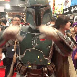 Video gif. A Boba Fett cosplayer forms a heart at us with his hands and tilts his head in a cute, loving way. 