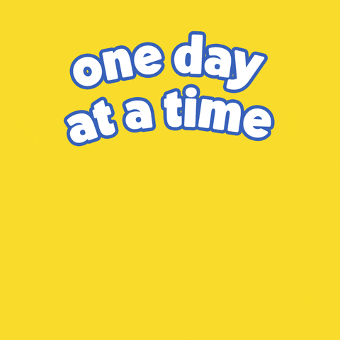 Digital art gif. Cartoon rainbow glides across the screen, a cheery smiley face sitting at the bottom arc of the rainbow. Big, white bubble text reads, "One day at a time," all against a bright yellow background.