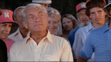 Movie gif. Ted Knight as Judge Elihu Smails in Caddyshack stands among a crowd looking smug and impatient, saying, "well, we're waiting."