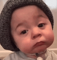 Video gif. A baby wearing a gray beanie stares at us with a sad expression on his face before slowly looking down remorsefully. 