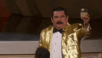 Oscars 2024 GIF. Guillermo holds up a tequila margarita and faces the audience. He's wearing a gold sequined tuxedo jacket. He cheers the crowd before clinking glasses with an audience member.